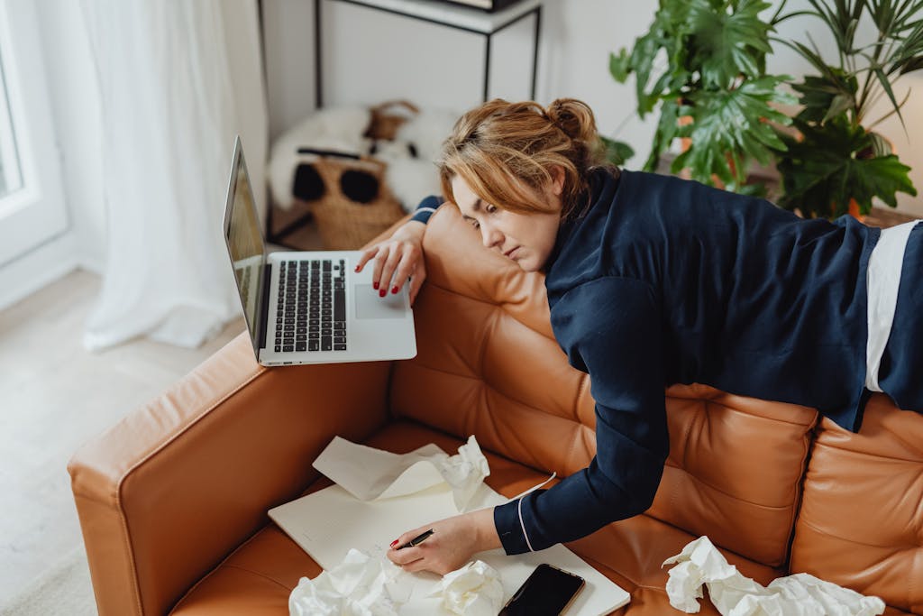 Woman resting on a couch with laptop, surrounded by papers, depicting stressed remote work lifestyle.